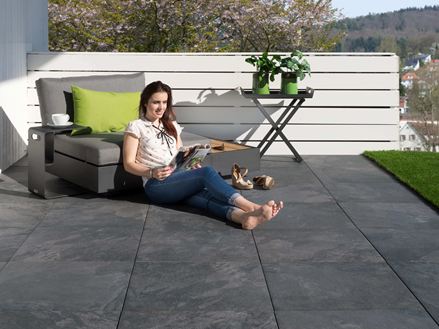 Młoda kobieta czyta na tarasie wyłożonym ciemnymi płytkami z gresu porcelanowego imitującymi łupek, wyposażonym w nowoczesne meble
